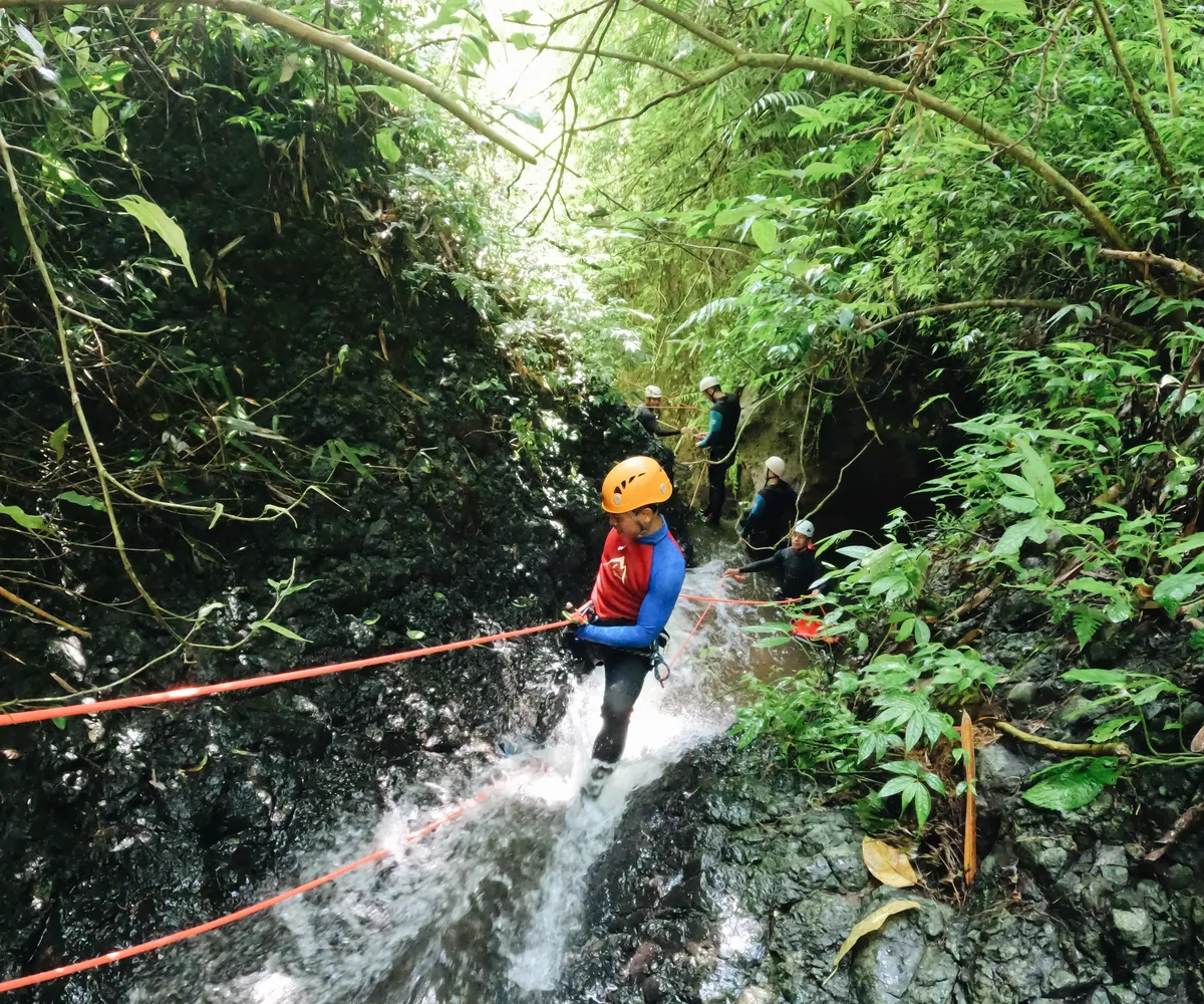 Canyoning participant in a helmet descending a waterfall in a green forest gorge