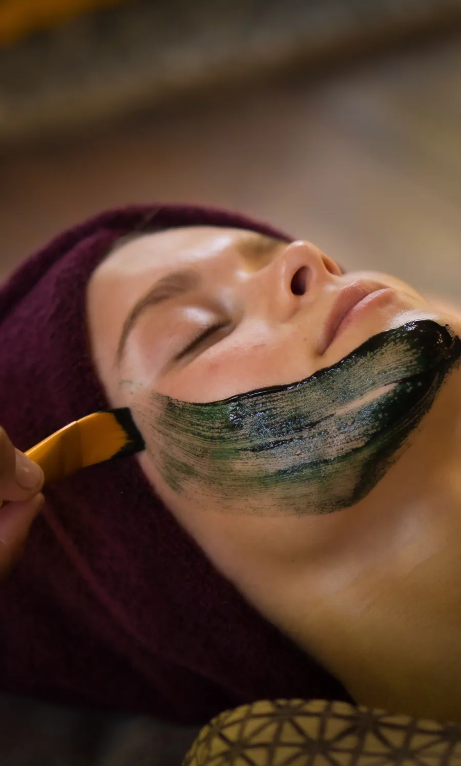 Guest in a maroon head towel receiving a facial treatment with a dark mask
