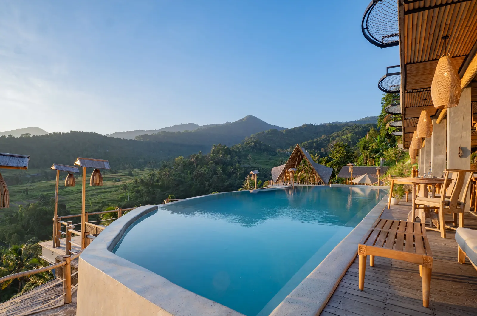 Infinity pool beside a timber terrace with woven lanterns, looking over forested hills under a clear blue sky