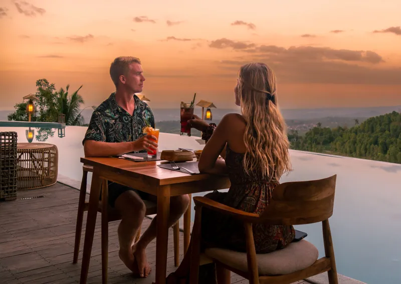 Couple having drinks at a table on a terrace beside an infinity pool at sunset
