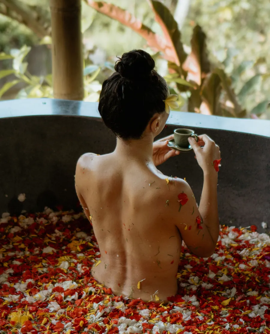 Woman seen from behind in a bath of flower petals, holding a small cup