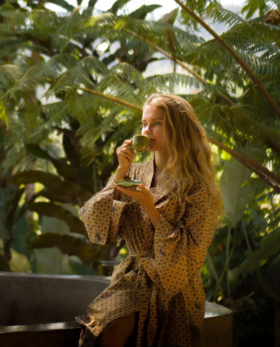 Woman in a patterned robe drinking tea among tropical foliage