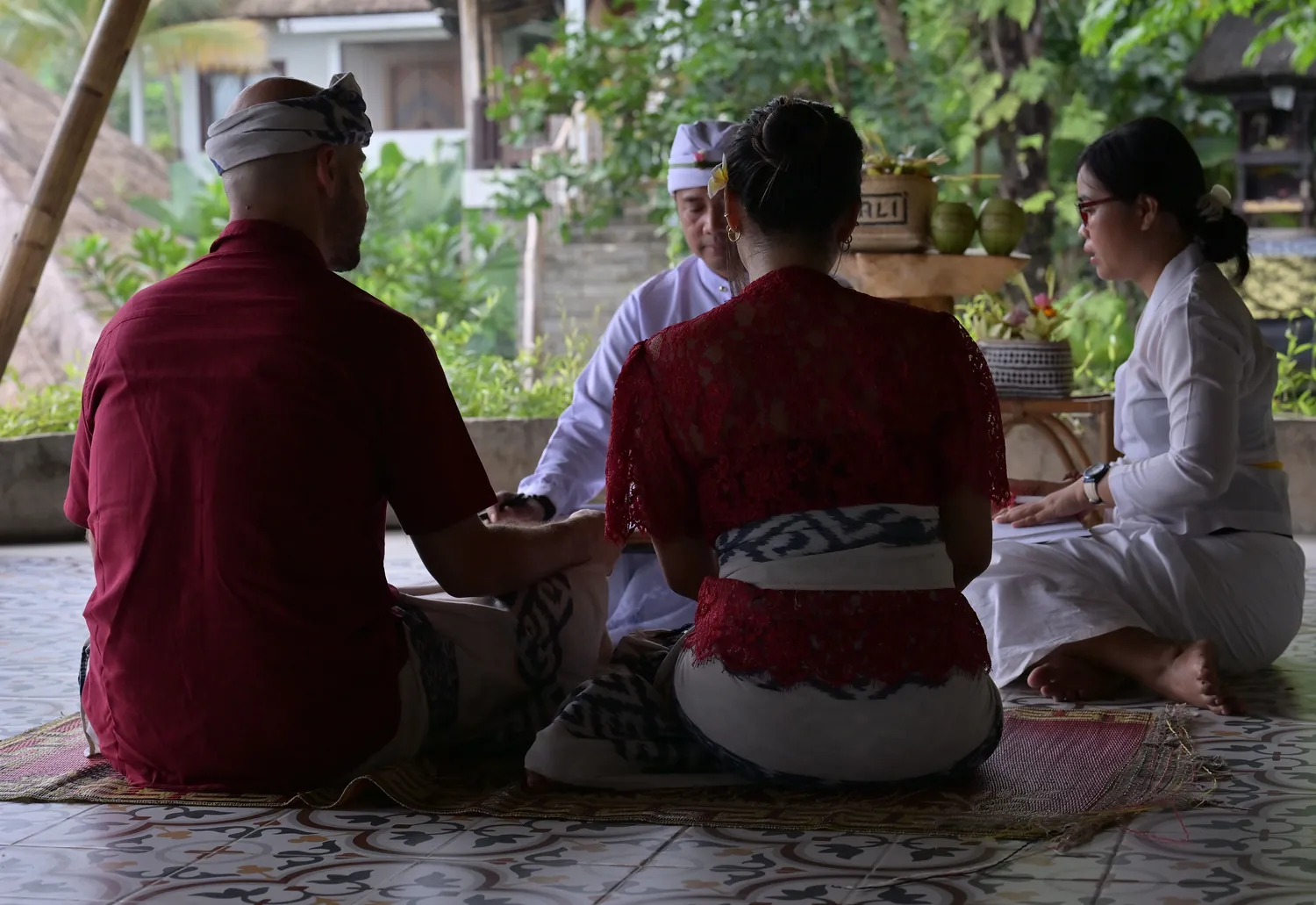 Two guests in red clothing seen from behind, seated on a woven mat facing two people in white