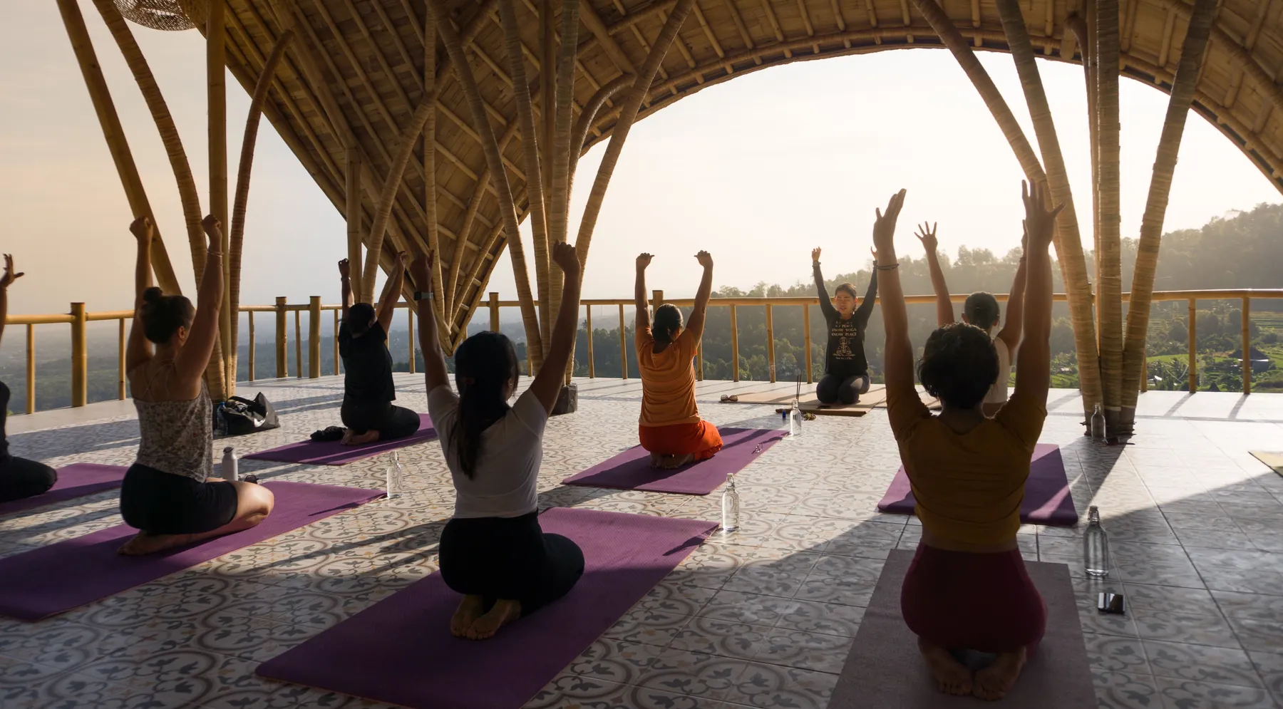 Guests in a yoga class with raised arms on purple mats under a bamboo roof, overlooking the hills at sunset